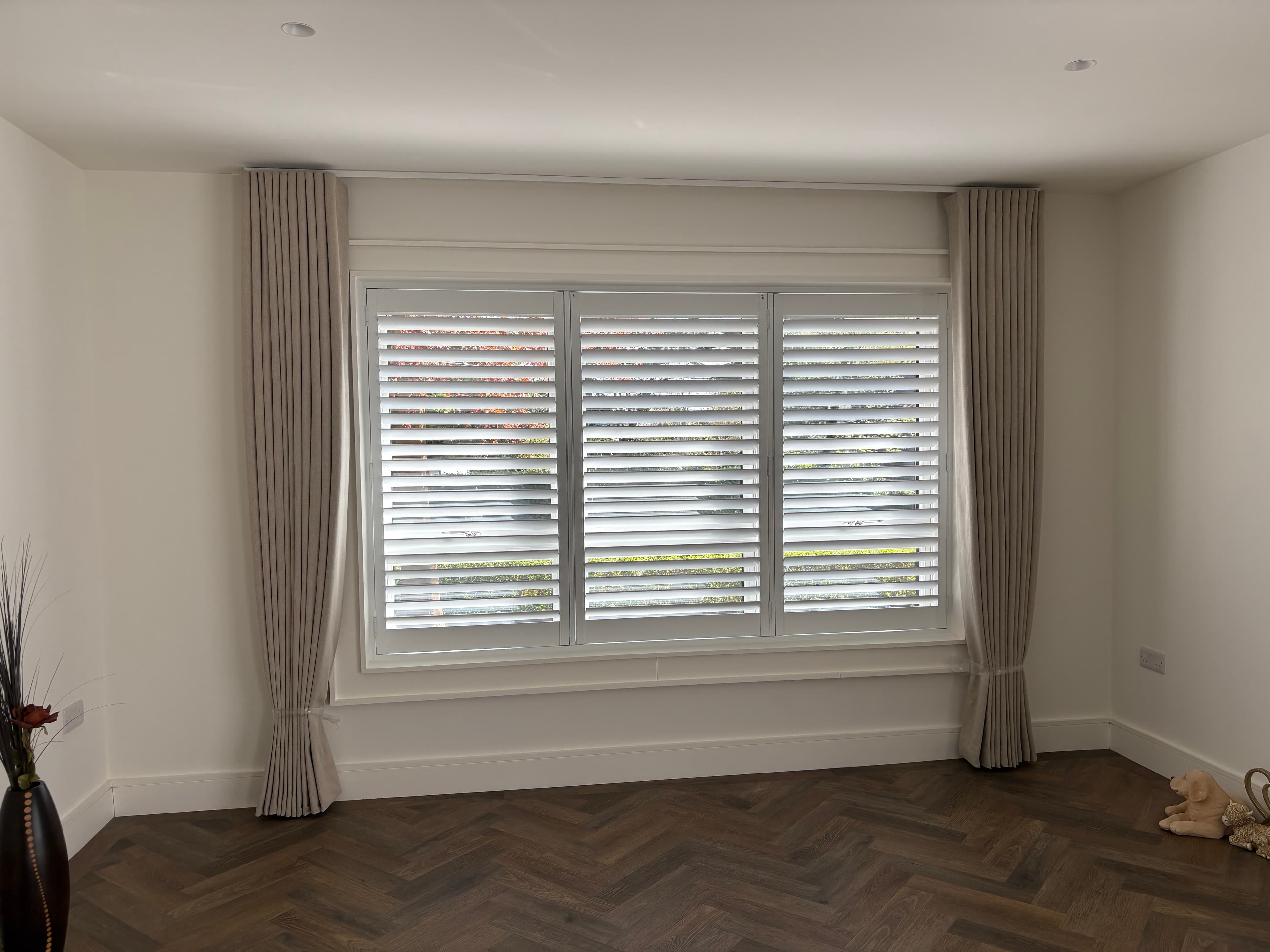 Three-panel white shutters framed by linen curtains on a track above a herringbone floor